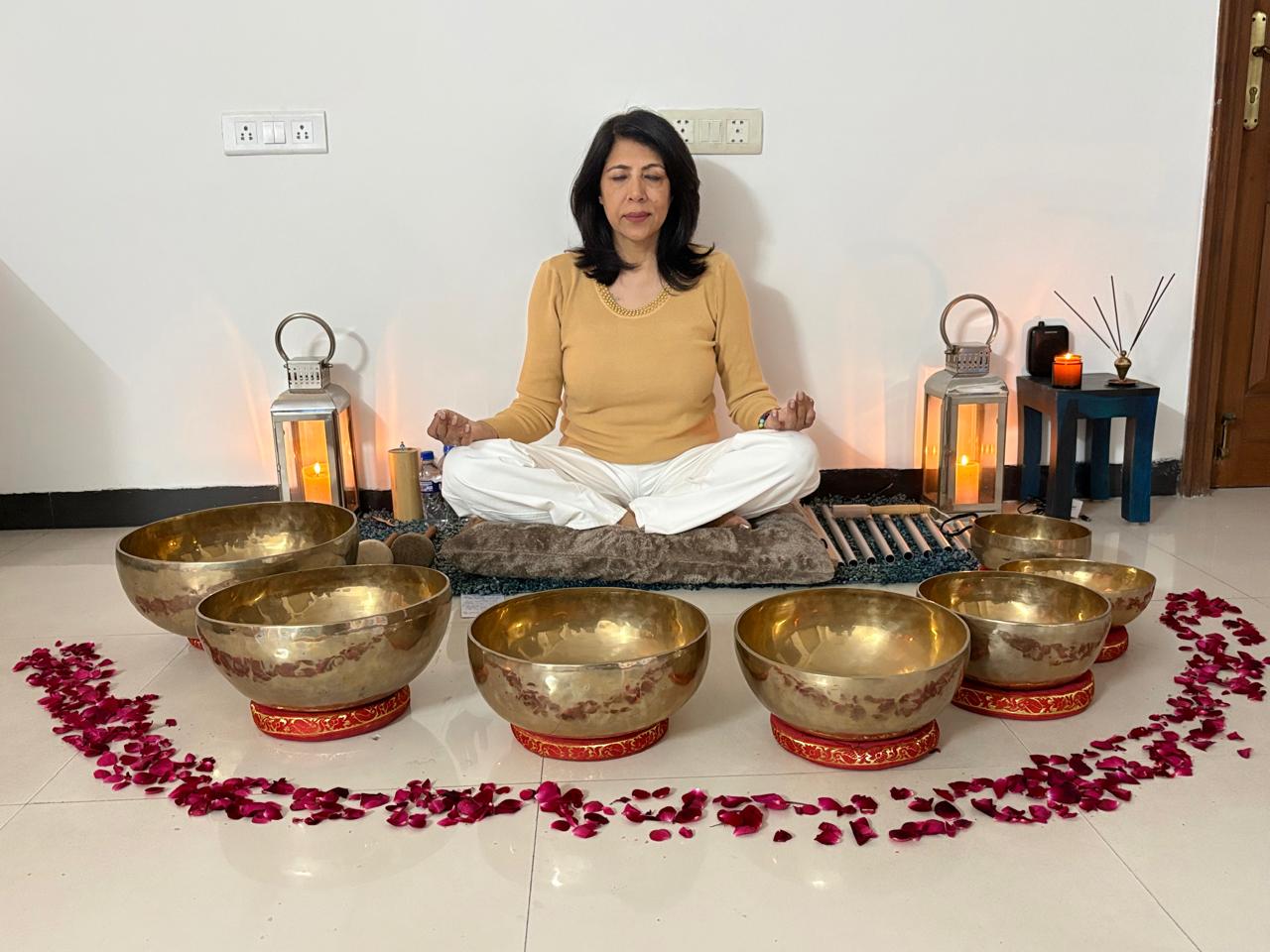 Shelly Rikhy Mandhar meditating peacefully with Tibetan singing bowls arranged in front of her, surrounded by rose petals and warm candlelight